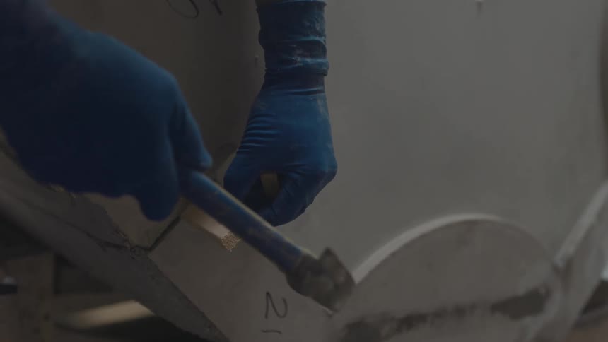 Gloved hands firmly grip a hammer as it strikes along the edge of a curved mold or surface in a dim industrial workspace