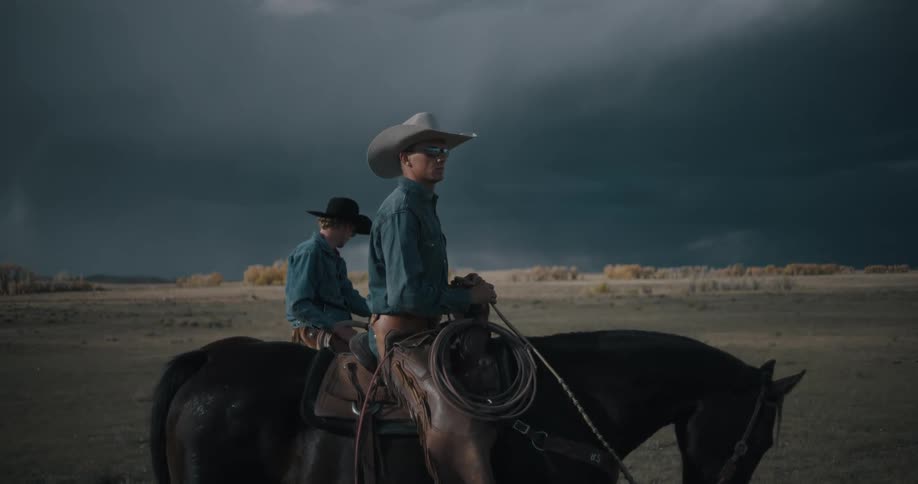 Two cowboys wearing hats and denim shirts sit atop their horses in a vast field looking out toward a horizon dominated by massive dark storm clouds