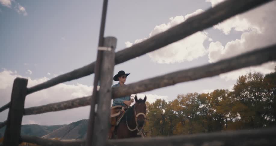 A cowboy in a widebrimmed hat and sunglasses rides a brown horse through a rustic dirt corral filmed from a low angle behind a wooden fence with vibrant autumn trees and mountains visible under a bright cloudy sky