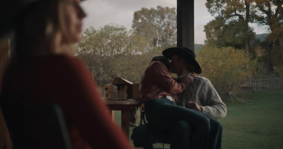 Couple kisses while seated on a porch as a blurred person appears in the foreground