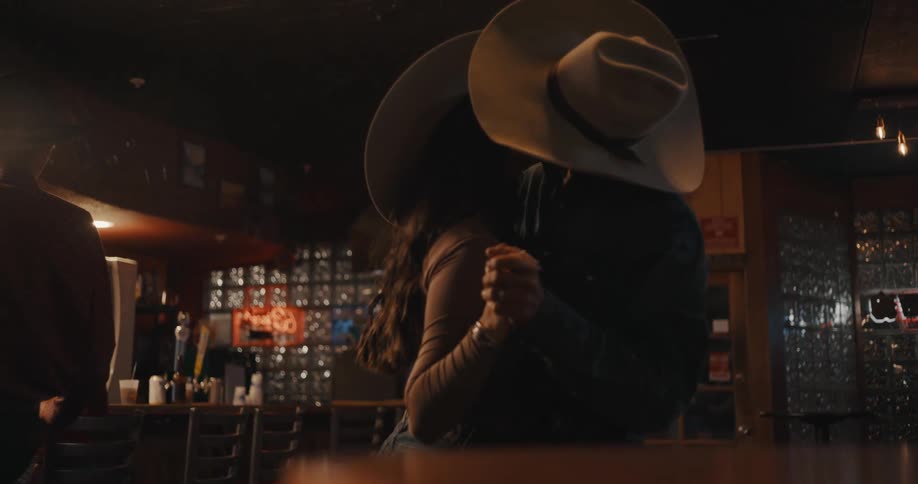 A man and woman wearing cowboy hats slow dance together in a dimly lit western bar rotating slowly while other persons sit at the counter in the background