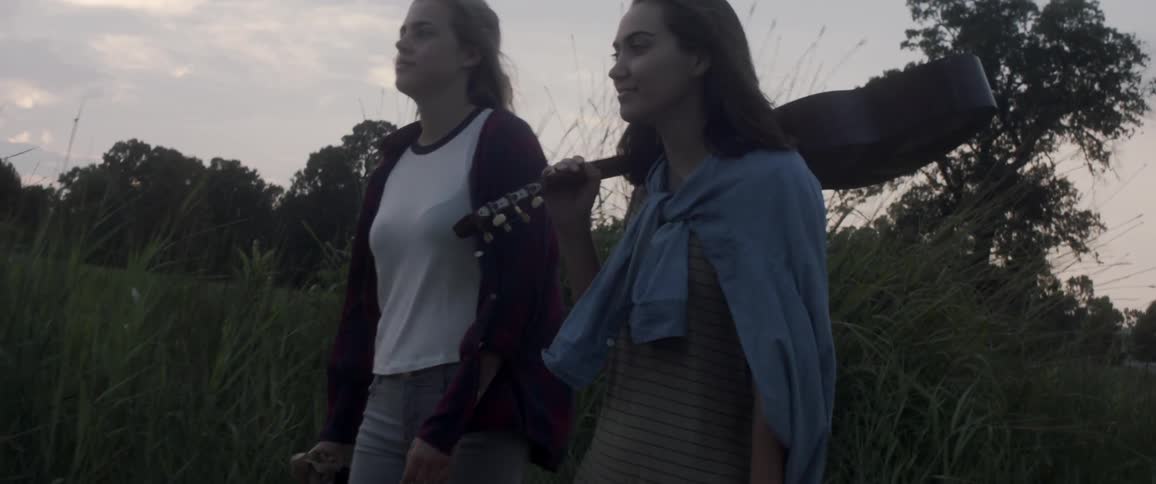 Two woman walk through tall grass and one is carrying a guitar