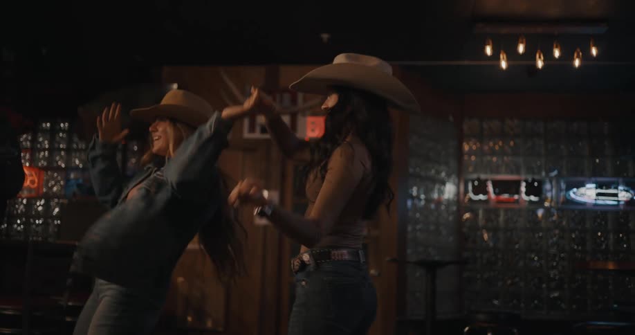 Women dance in a bar wearing cowboy hats under low lighting