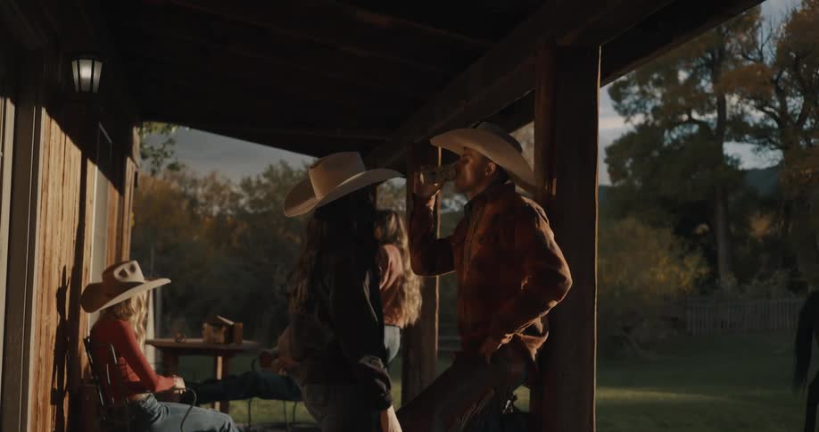 Group of people wearing cowboy hats talk and drink on a wooden porch at dusk