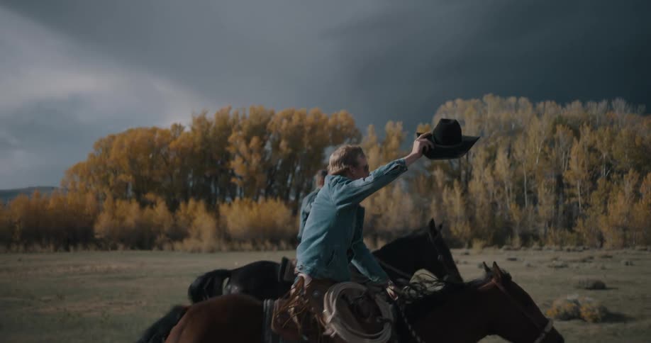 Riders on horseback raising hats in an open field with trees in the background and a heavy sky