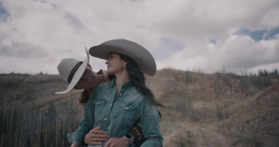 Couple wearing cowboy hats embracing in a rural area with hills in the background