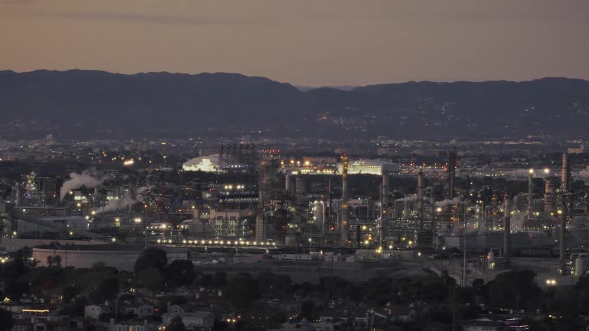 Industrial complex illuminated at dusk with an urban area and mountains in the background