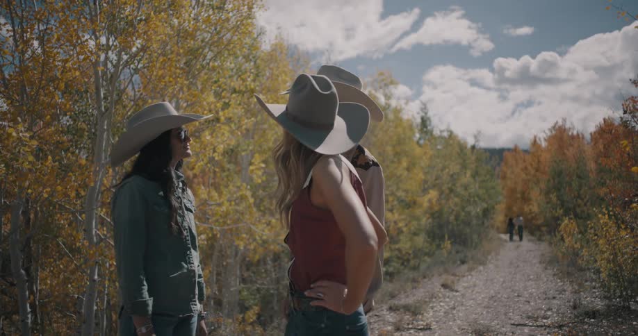 Three people wearing cowboy hats stand on a dirt path and talk together surrounded by yellow autumn trees and mountains under a bright sunny sky