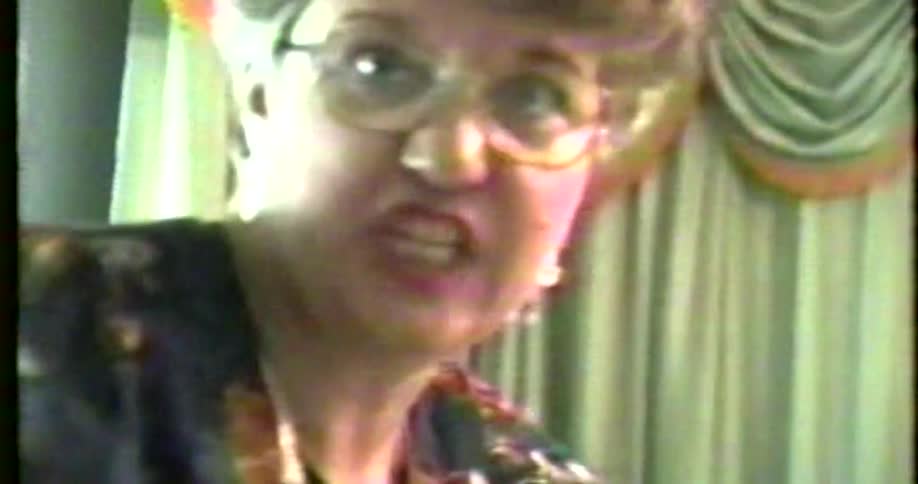 Adult woman wearing glasses and jewelry talking with a nervous facial expression and then smiling while cutting a celebration cake indoors