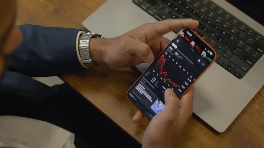 Man wearing a jacket checking price graphs on a mobile phone with a laptop comptuter in front of him on a wooden desk indoors