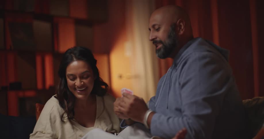 A man and woman sit on a sofa playing cards together the woman cheers and gives the man a highfive to celebrate her win while they enjoy wine and snacks in a dimly lit room