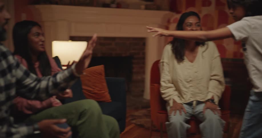 A group of family and friends share a joyful evening together laughing and playing games in a warm cozy living room one young woman expresses surprise and delight while holding up a small blue object during their festive gathering