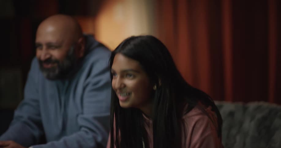 A teen and an older man sit in a living room smiling and celebrating as they look forward while the teen holds a game controller
