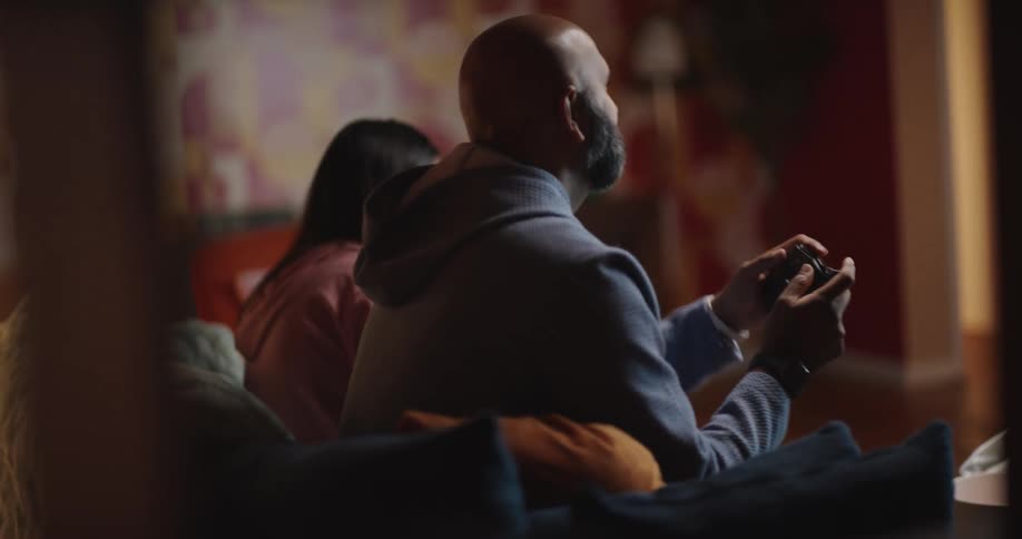 A man sits on a sofa next to a young woman and focuses on a television screen while holding a video game controller engaging in a quiet gaming session within a dimly lit and cozy living room during the evening
