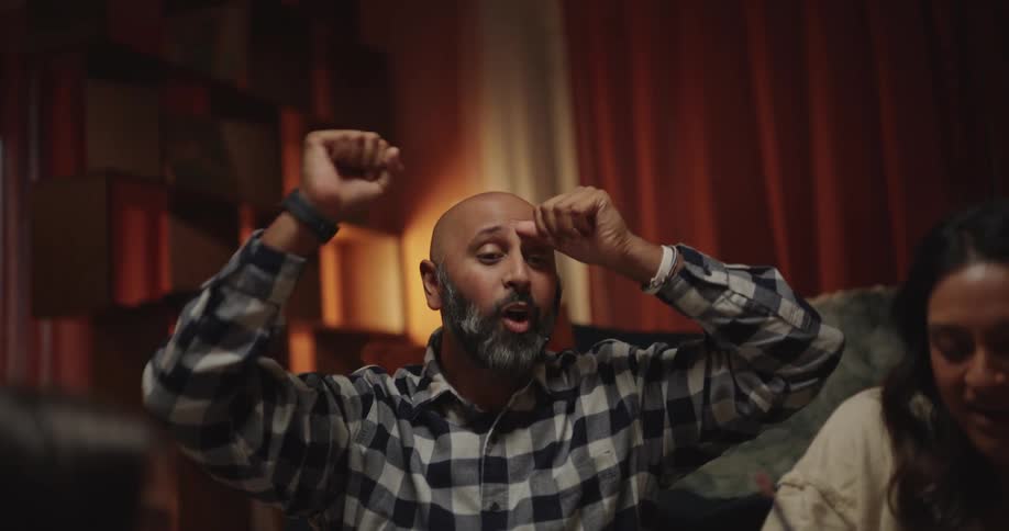 A man with a gray beard wearing a plaid shirt shakes a blue cup and celebrates with raised arms while playing a board game with a woman and two girls in a room with warm lighting