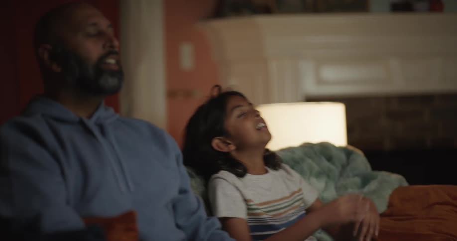 Father and daughter playing video games with a controller while sitting on a couch in a living room