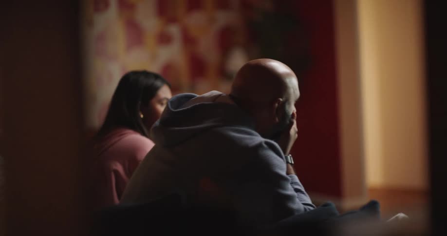 A man holding a game controller reacts with frustration as a woman sitting next to him jumps up in celebration the two sit together on a couch in a dimly lit room with patterned wallpaper experiencing contrasting emotions during their play