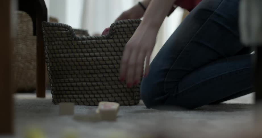A woman in a living room picking up toys and placing them in a basket