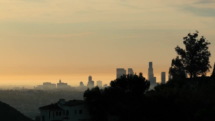 Video features a closer view of the same urban skyline shrouded in haze framed by dark trees and a hillside home at dusk