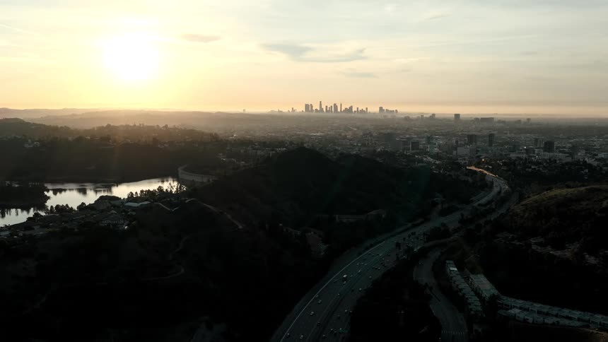 A stunning sunrise casts a golden glow over a sprawling city as a busy highway weaves through shadowy hills beside a tranquil reservoir