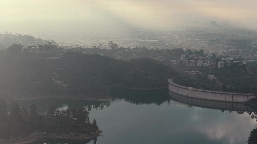 A high angle view of a calm lake and a curved dam wall with a vast city shrouded in thick morning haze and soft sunlight in the background
