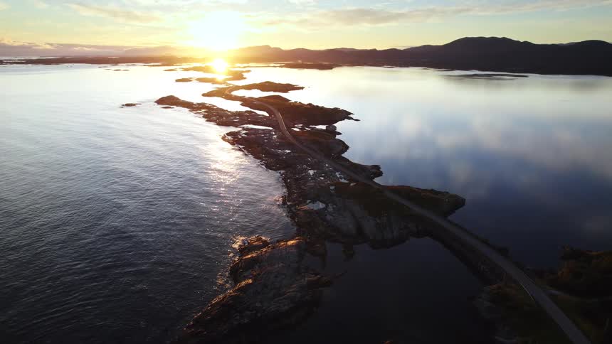 A high aerial view shows a coastal road winding across rocky islands surrounded by calm blue water as the sun sets over distant mountains