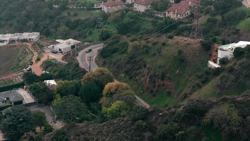 An aerial shot shows a winding road cutting through lush green hills dotted with houses and trees several cars drive along the asphalt path as the camera slowly pans over the scenic landscape