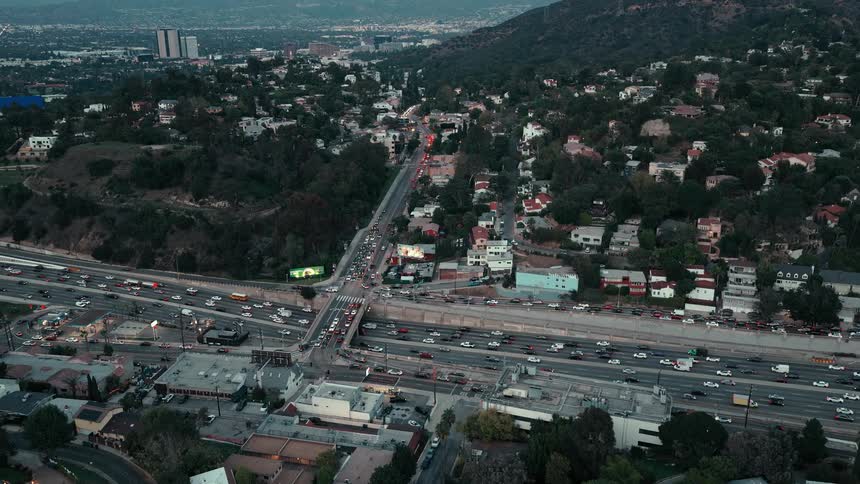 At dusk an aerial view captures the steady flow of heavy traffic along a major freeway nestled beside a hillside residential area as city lights glow in the distance