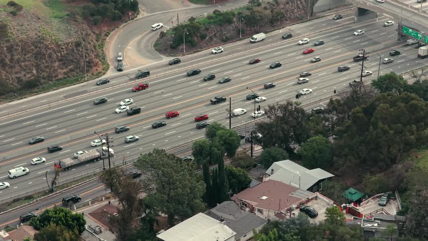 An aerial shot shows heavy traffic flowing along a multi lane freeway that cuts through a hilly residential area