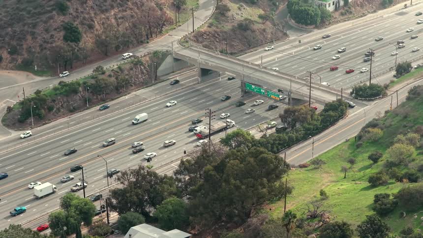 This aerial shot captures a busy highway with multiple lanes of traffic flowing steadily through a hilly landscape a bridge spans across the freeway while lush greenery and residential buildings dot the surrounding terrain