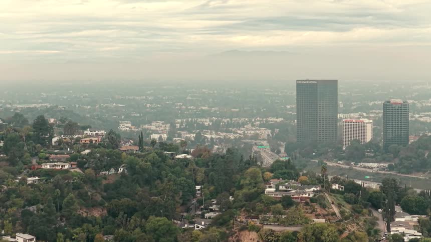 This aerial shot captures a hazy cityscape with distant mountains partially obscured by a thick layer of smog the camera pans slowly over lush residential hills in the foreground showcasing homes nestled among dense green trees