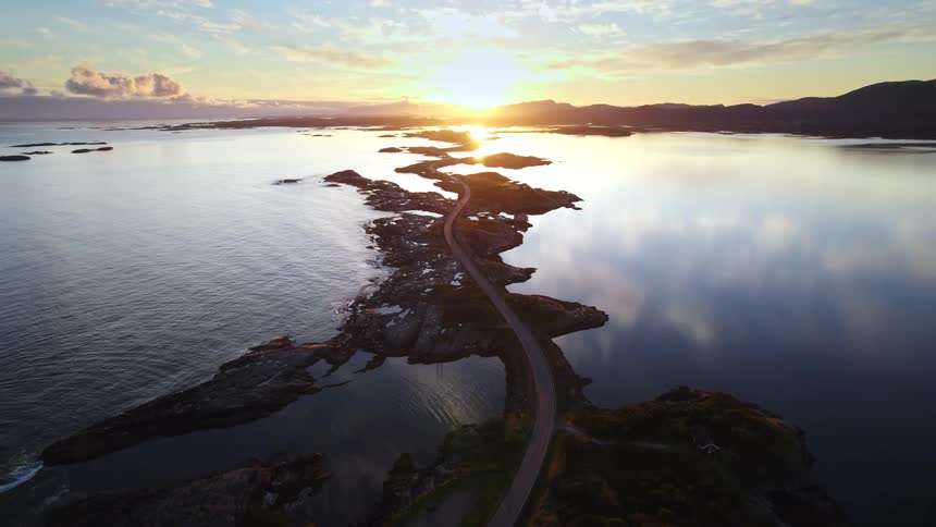 An aerial view captures the atlantic ocean road in norway at sunset where a narrow highway curves across small rocky islands and bridges the golden sun reflects brilliantly on the calm mirrorlike water surrounding the jagged coastline
