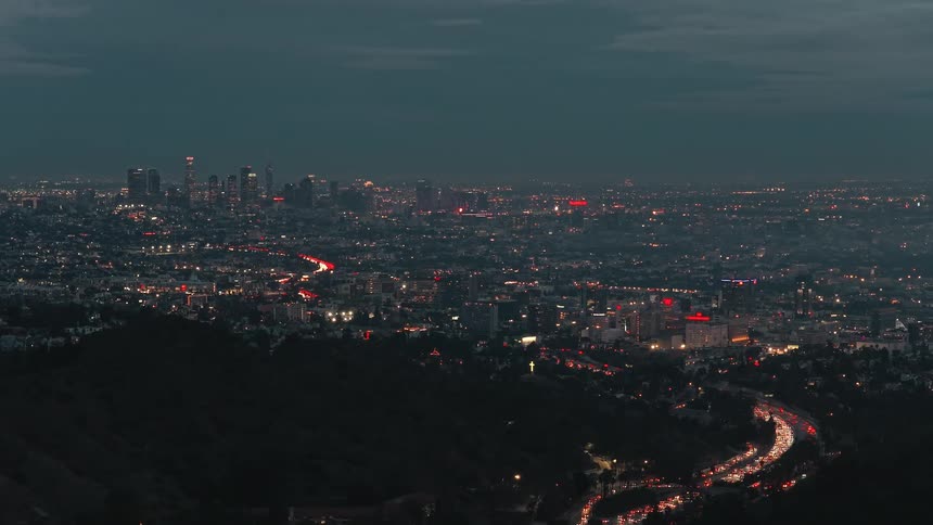 A wide elevated view captures a sprawling city at dusk as thousands of lights flicker across the urban landscape and long lines of traffic flow steadily along winding highways toward a distant cluster of illuminated skyscrapers