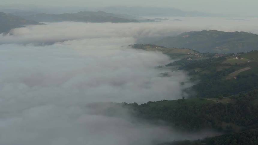 An aerial wide shot shows a thick blanket of white fog settling into deep mountain valleys obscuring the ground while dark green forested ridges and small patches of developed hillside emerge through the moving mist under a pale sky