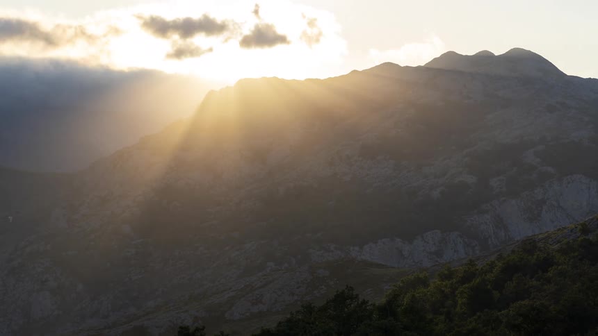 Rocky mountain with another mountain covered in trees next to it and the sun in the sky in the background during dusk or dawn
