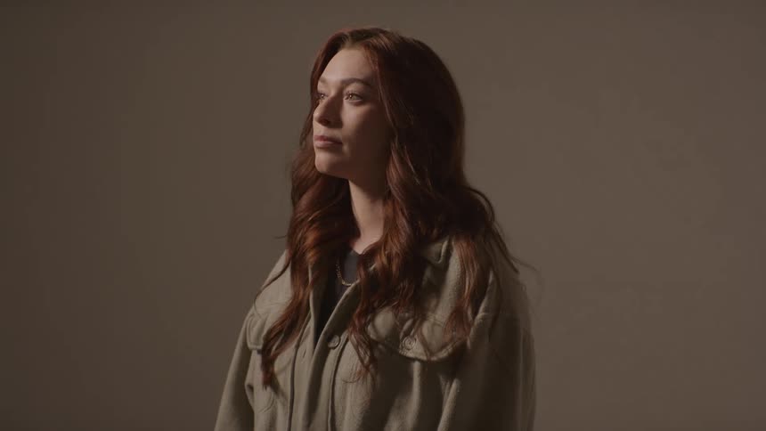 Person with wavy auburn hair standing against neutral background with soft lighting