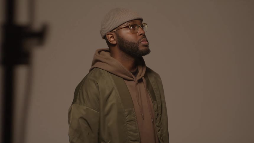 A man wearing a beanie glasses and a jacket stands in a studio looking slightly upward as shadows shift across his face and the warmtoned background