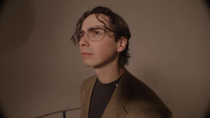 A man wearing glasses and a tweed jacket stands in a dimly lit room his expression shifting from pensive to serious as he looks off camera