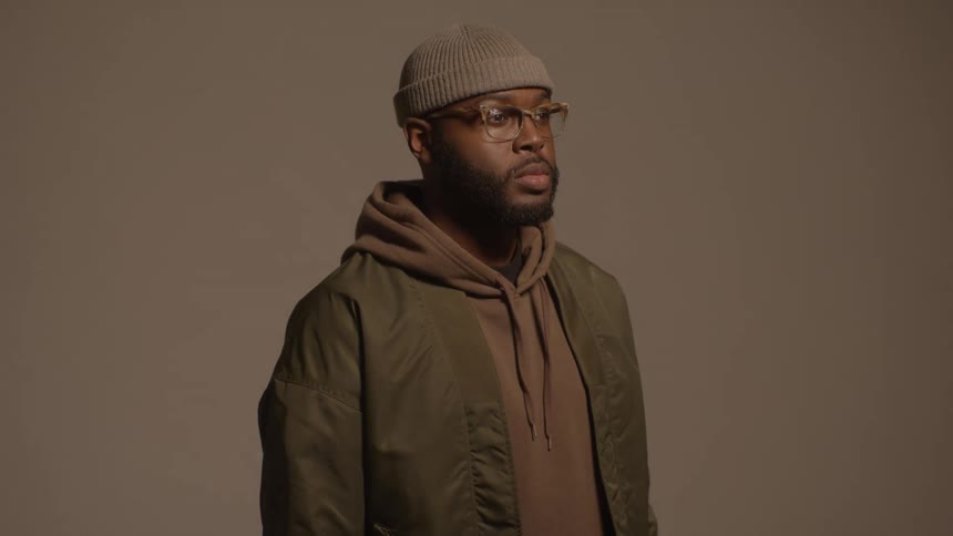 A man wearing a beanie glasses and a layered jacket stands against a neutral background while looking off to the sidethe lighting gradually brightens across his face throughout the clip revealing more detail in his expression and clothing