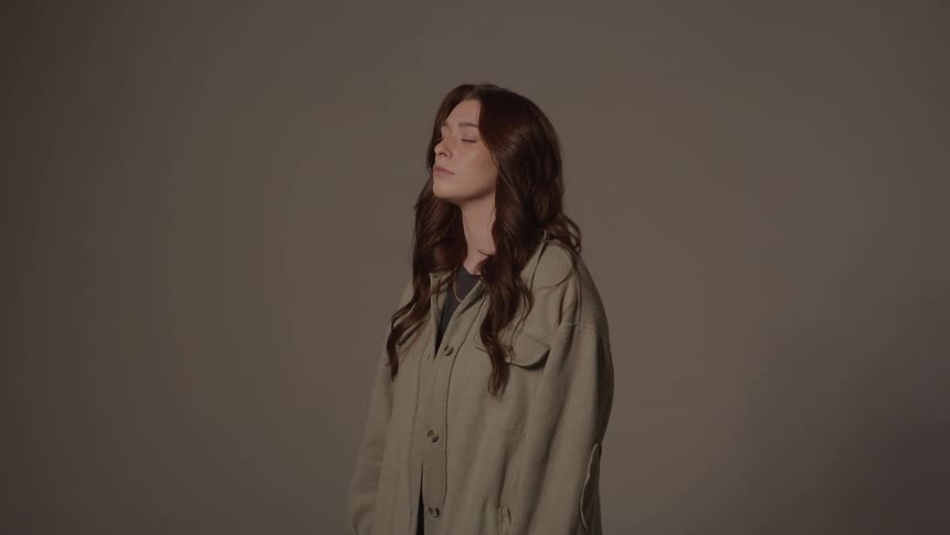 Woman with closed eyes and wavy brown hair standing against neutral background