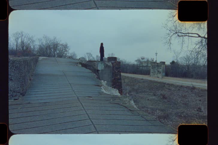 A person in a dark coat with a red hood walks onto a decaying bridge in a desolate overcast landscape and sits down