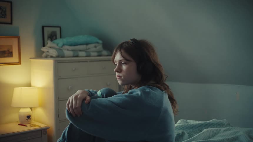 A young woman sits alone on her bed in a dimly lit room hugging her knees with a somber expressionshe gazes off into the distance appearing lost in deep and troubled thought while the warm glow of a bedside lamp contrasts with the cool tones of the r
