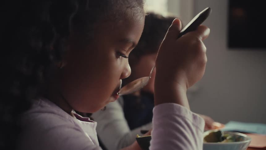 Two young girls sit side by side at a table enjoying a meal togetherone girl in the foreground takes a large spoonful of food while her companion eats in the background