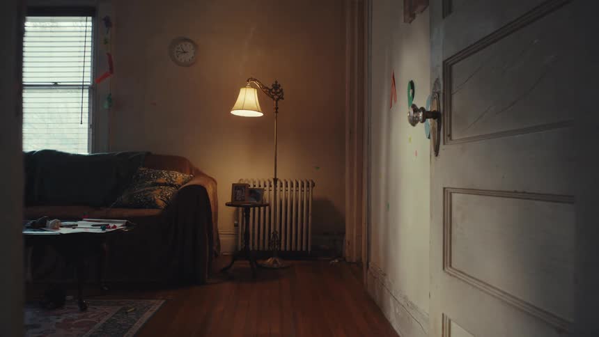 A static wide shot shows a dimly lit empty living room with hardwood floors featuring a brown sofa near a window with closed blinds a glowing floor lamp beside a radiator and an open white door in the foreground