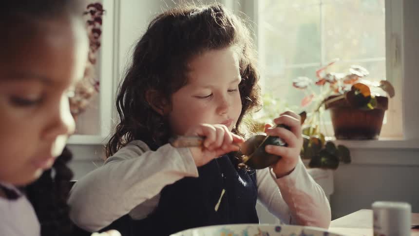 Two young girls sit at a dining table bathed in natural light with one intently scooping out and eating an avocado with a spoon