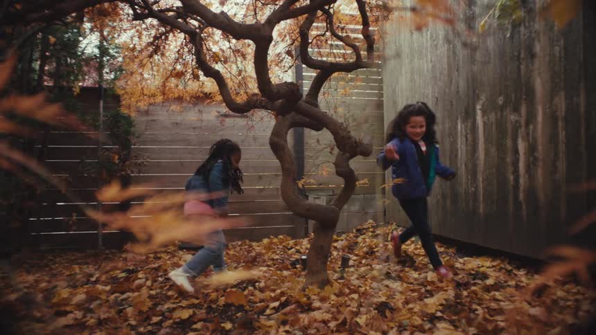 Two girls running and playing around trees near a fence where the ground is covered with fallen leaves in bright autumn colors