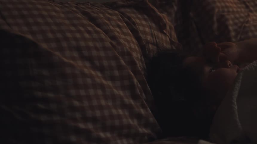 In a dimly lit cozy bed a young child wakes up rubs their eyes and then settles back down to sleep