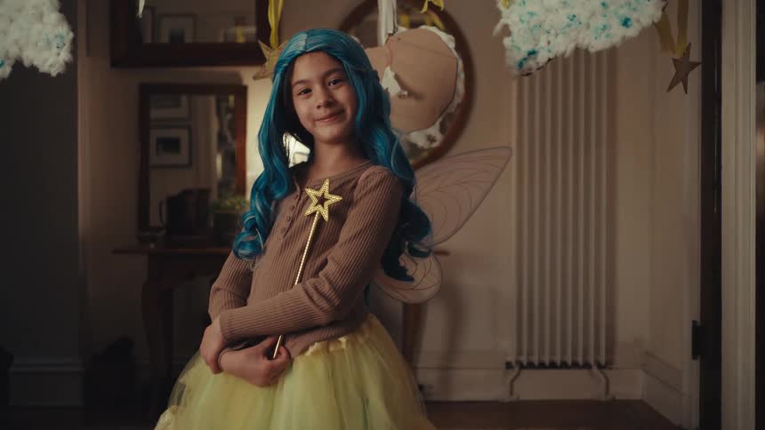 A young girl wearing a blue wig fairy wings and a yellow tutu stands in a hallway holding a gold star wand while looking directly into the camera with a slight smile as decorative clouds hang from the ceiling around her