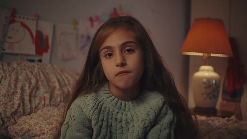 Child with long brown hair in light blue sweater seated in home setting with childrens drawings on wall