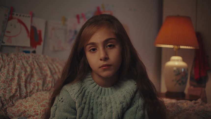 Little girl sitting on a bed in a room with a lamp in the background